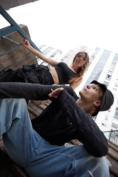 Low angle view of a teenage couple poses in the urban exterior on the stairs surrounded by skyscrapers and buildings.