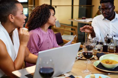 Multicultural colleagues sit in a restaurant at business lunch while listening to their boss explaining the project.