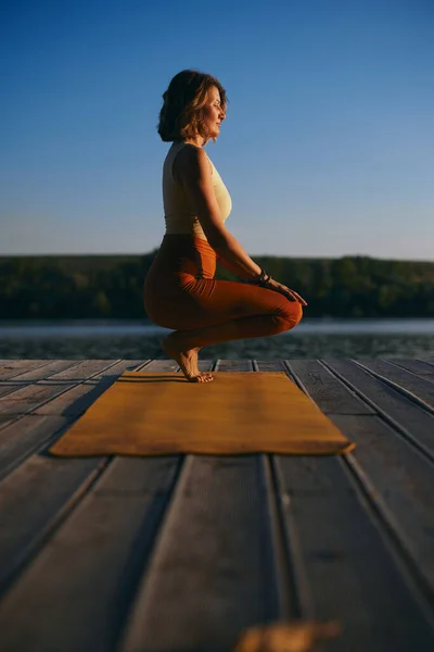 The tranquil woman is practicing yoga on the dock while crouching and balancing. She is in the Squatting Toe Balance yoga pose.