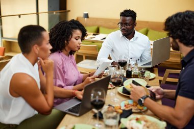 Multicultural colleagues sit in a restaurant at business lunch while listening to their boss explaining the project.