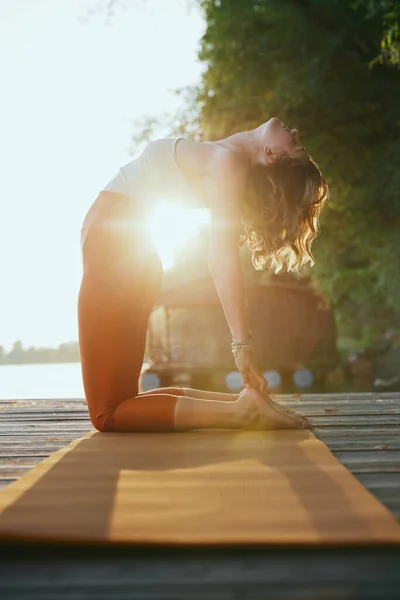 A fit middle-aged woman is practicing yoga on the dock at sunset. She is in the Camel yoga pose.