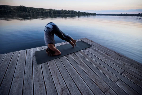 A fit woman practices yoga on the dock near the river and balances on her head in the Supported Headstand yoga pose.