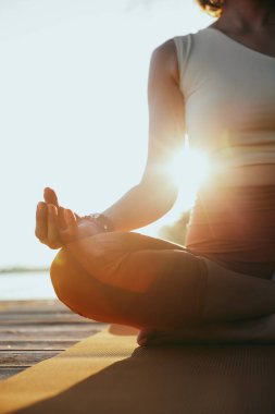 A calm woman sits on the dock in a lotus pose and meditates.