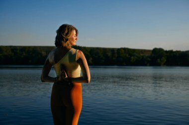 Back view of yogi woman holding hands in namaste pose and meditating on the dock near the lake.