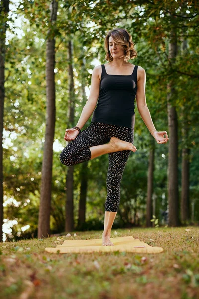 A slim woman practices yoga in the forest in autumn. She is in the Standing Half Lotus yoga pose.