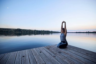 A calm yogi woman in a lotus position is meditating on the dock.
