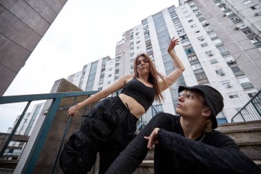 A teenage couple is posing outside on the stairs surrounded by buildings.