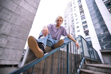 A teenage boy is sitting in an urban exterior surrounded by buildings.