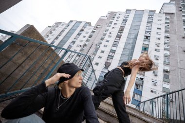 A teenage couple is posing outside on the stairs surrounded by buildings.