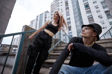 A teenage couple is posing outside on the stairs surrounded by buildings.