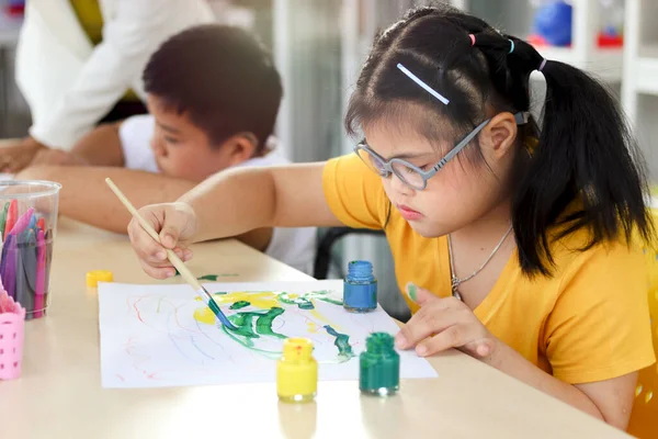 Happy children having fun with friends during study at school, girl with down syndrome concentrate painting on paper in art classroom, education of kids with physical disability and intellectual concept.