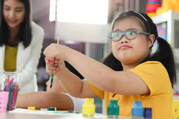 Happy children having fun with friends during study at school, girl with down syndrome concentrate painting on paper in art classroom, education of kids with physical disability and intellectual concept.