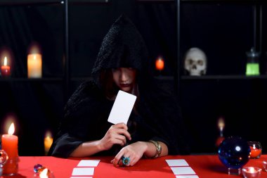 Siyah başlıklı kara cadı elinde sihirli kart, gizemli güzel kadın falcı geleceği kartla okuyor, korkutucu Cadılar Bayramı konsepti..