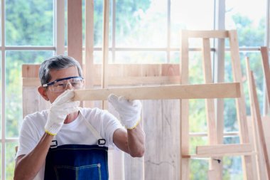 Güvenlik gözlüğü ve bez eldiven takan yaşlı marangoz, ahşap kalas tutuyor ve nişan alıyor, marangozluk atölyesindeki gözleriyle kereste uzunluğunu tahmin eden usta zanaatkar, DIY hobisinin keyfini çıkarın..