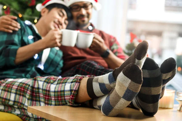 Lover feet in socks on table, relaxing adorable LGBT couple sharing special moment together on Christmas holiday, Asian gay male sitting on sofa, drinking hot beverage chocolate to celebrate Christmas