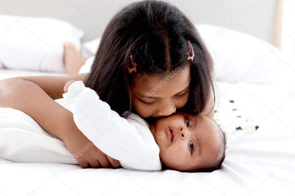 A little Asian African newborn infant baby girl lying on white bed with her sister who hugging and kissing her, sweet adorable kid showing love and care to family members