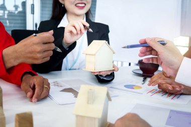 İş adamları ve mühendis toplantı masasında inşaat planını tartışıyor, kalemli ahşap bir ev modelini işaret ediyor, ofisteki inşaat projesinde ekip birlikte konuşuyor..