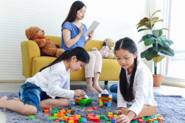 İki kız çocuğu tahta blok oyunu (Jenga) ile oynarken, anne kanepede oturup internette sörf yapıyor, anne tablet kullanarak çalışıyor, kız kardeş çocuk birlikte vakit geçiriyor..