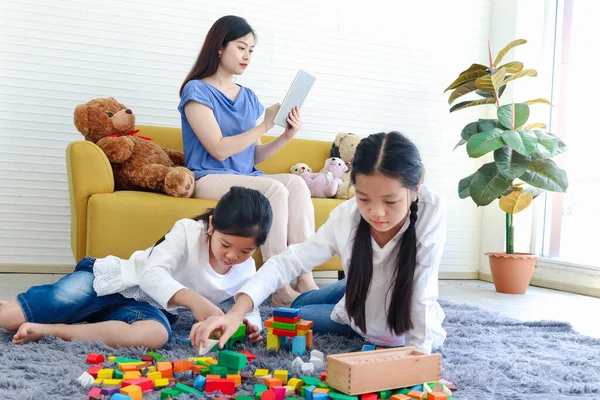 İki kız çocuğu tahta blok oyunu (Jenga) ile oynarken, anne kanepede oturup internette sörf yapıyor, anne tablet kullanarak çalışıyor, kız kardeş çocuk birlikte vakit geçiriyor..