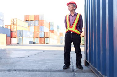 Güvenlik yeleği ve miğfer giyen yaşlı Asyalı işçi mühendisinin portresi lojistik kargo konteynırları deposunda duruyor..