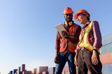 Güvenlik yeleği ve kask giyen iki işçi lojistik kargo konteynır deposunda tartışmak için dijital tablet kullanıyorlar. Afrikalı Amerikalı ve kıdemli yaşlı Asyalı mühendis birlikte çalışıyorlar.