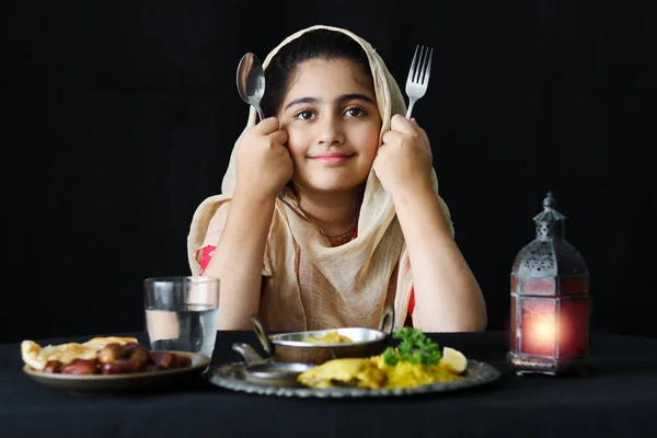 キッチンテーブルに座ってスプーンやフォークを持って暗い黒の背景にイスラム教徒のハラル料理を持っているための美しい目を持つ愛らしい笑顔パキスタンのイスラム教徒の女の子 — ストック写真
