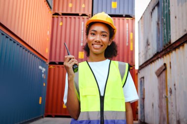 Mutlu gülümseyen, kıvırcık saçlı, çelik yelek ve miğfer giyen, lojistik kargo konteynır deposunda telsiz taşıyan, güzel, Afro-Amerikan kadın mühendis portresi..