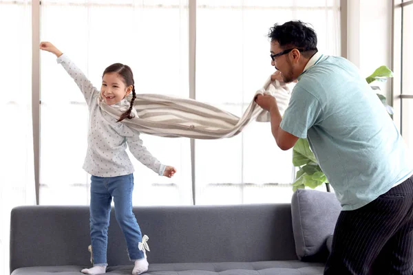 可爱的小女孩穿着大毛巾 穿着超级英雄的衣服 父亲一边弹着布 一边假装她能在客厅的沙发上飞翔 孩子和爸爸一起在家里扮演英雄 父亲节快乐 — 图库照片