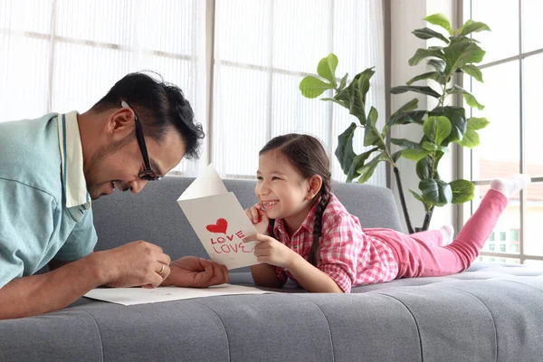 父亲节快乐 可爱可爱的小女孩和年轻的爸爸躺在客厅的沙发上 手工制作贺卡进行校准 快乐的亲生父母在一起 — 图库照片