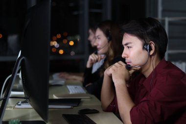 Mutlu gülümseyen Asyalı adam gece yarısı takım arkadaşlarıyla çağrı merkezinde masa başı danışmanı, mikrofon kulaklığı olan müşteri hizmetleri yöneticisi gece geç saatlerde ofis desteği için bilgisayar kullanıyor..