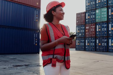 Kıvırcık saçlı, güvenlik yeleği ve miğferi olan Afro-Amerikan mühendis kadın mühendis. Alacakaranlık saatinde lojistik kargo konteynerinde cep telefonu tutuyor..