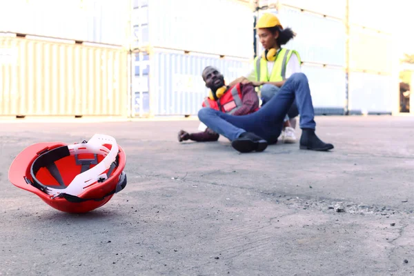 Safety helmet on ground with background of industrial African American engineer worker man getting hurt from accident at workplace, lying on floor at construction site area, colleague try to help him.