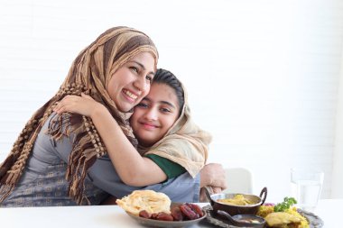 Sevimli Pakistanlı Müslüman kız geleneksel İslami helal yiyeceklerle mutfak masasında oturuyor, tesettürlü çocuk annesine sarılıyor, anne ve kızına karşı sıcak bir sevgi besliyor, beyaz arka planda mutlu bir İslami aile..