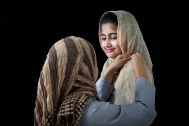 Pakistanlı Müslüman anne kızını geleneksel başörtüsü ile giydiriyor, güzel gözlü sevimli çocuk annesine gülümsüyor, koyu siyah arka planda mutlu bir İslami aile..