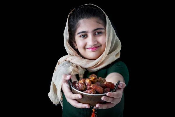 美しい目を持つ愛らしい笑顔パキスタンのイスラム教徒の少女は カメラに向かって日付の果物を保持ヒジャーブを身に着けています 甘さとエネルギーは闇の黒を背景にイスラム教のラマダーン月の食事を促進します — ストック写真
