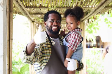 Mutlu çiftçi ailesi tarım çiftliğinde birlikte çalışıyor, gülümseyen Afrikalı baba siyah kıvırcık saçlı küçük kızını tutuyor ve kameraya, çocuğa ve hediyelere parmağını uzatarak bahçenin tadını çıkarıyor..