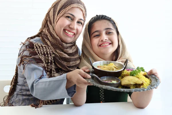 Adorable smiling Pakistani Muslim girl and mother at kitchen, hold tray of traditional Islamic food, daughter and mom wear hijab, show halal food to camera, happy Islamic family on white background.