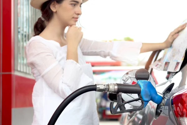 Blue fuel petrol pump nozzle against refueling car with petrol with beautiful female traveler holding touristic map and waiting as blurred background at gas station, customer woman self-refueling car.