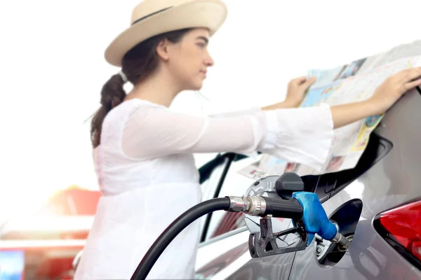 Blue fuel petrol pump nozzle against refueling car with petrol with beautiful female traveler holding touristic map and waiting as blurred background at gas station, customer woman self-refueling car.