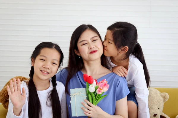 Two daughter girls give tulip flower and present gift box for smiling mother while hug and kiss mom with love at living room, warm love in family. Happy mother day, children have special time with mom