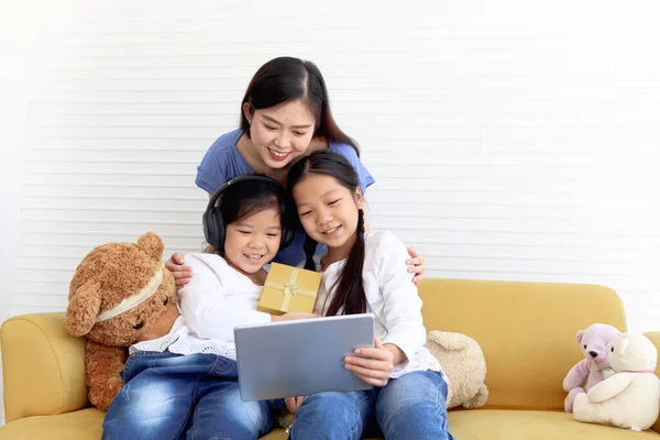 Asia happy family, cute two daughter girls with mother hold digital tablet, sister give present gift box to camera on video call, smiling mom and children have fun meeting online, surfing internet.