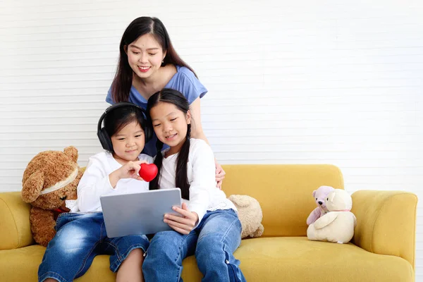 Adorable Asia happy family, cute two daughter girls with mother hold digital tablet, look and give red heart to web camera on video call, smiling mom and children have fun meeting online.