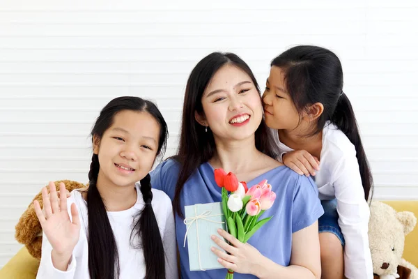 Two daughter girls give tulip flower and present gift box for smiling mother while hug and kiss mom with love at living room, warm love in family. Happy mother day, children have special time with mom