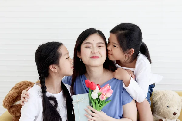 Two daughter girls give tulip flower and present gift box for smiling mother while hug and kiss mom with love at living room, warm love in family. Happy mother day, children have special time with mom