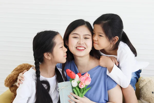 Two daughter girls give tulip flower and present gift box for smiling mother while hug and kiss mom with love at living room, warm love in family. Happy mother day, children have special time with mom