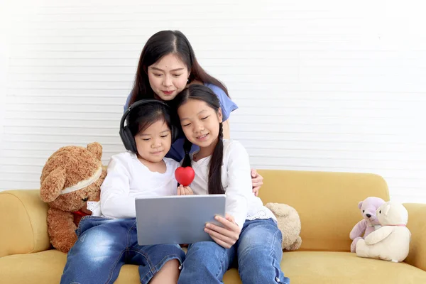 Adorable Asia happy family, cute two daughter girls with mother hold digital tablet, look and give red heart to web camera on video call, smiling mom and children have fun meeting online.