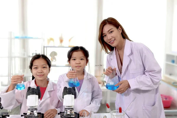 Students and teacher in lab coat have fun together while learn science experiment in laboratory. Young pretty Asian scientist and schoolgirl kids showing blue chemical flasks, little children students study research at school lab with female teacher.