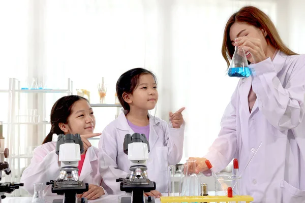Students and teacher in lab coat having fun together while learning science experiment in laboratory. Young pretty Asian scientist showing chemical flasks to two adorable schoolgirls and girls pointing, little children students study research at scho