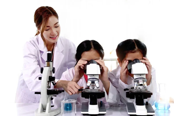 Students and teacher in lab coat have fun together, two little children students look through microscope and learn science experiment in laboratory with female teacher in classroom. Young pretty Asian scientist helps schoolgirl kids use lab equipment
