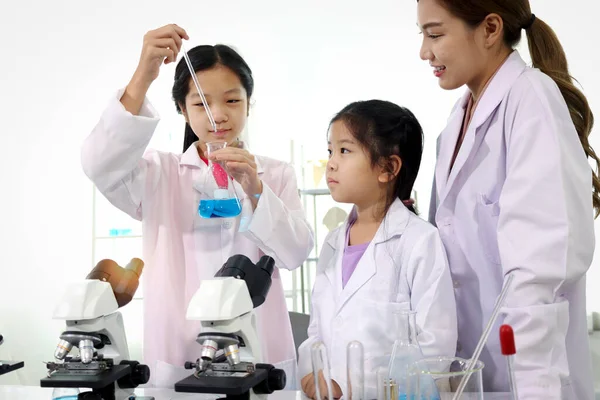 Students and teacher in lab coat have fun together while learn science experiment in laboratory. Young adorable Asian scientist kid adding some chemical to blue flask with pipette, little children students study research at school lab. Science educat